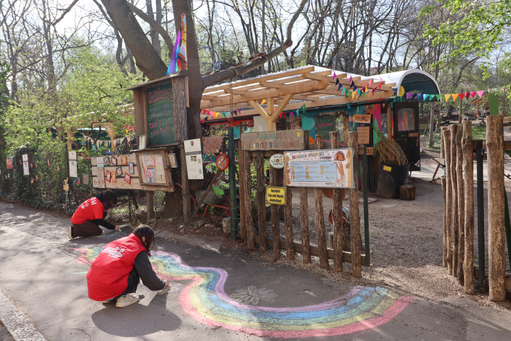 Zwei Kinder malen einen Regenbogen auf dem Gehweg. Im Hintergrund sieht man einen Park mit einem umgebauten Eisenbahnwagon.