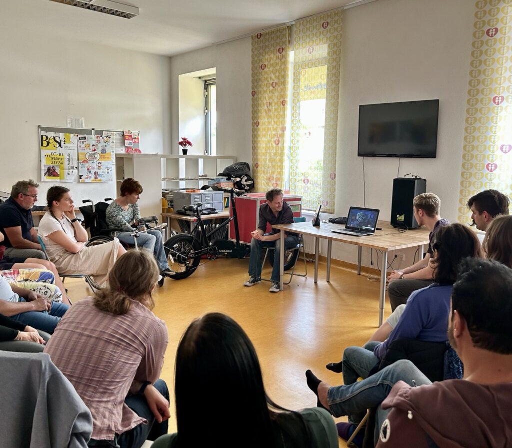 Eine Gruppe von Menschen sitzen in einem Raum im Kreis um einen Tisch und schauen auf einen Laptop, der aufgeklappt auf dem Tisch steht. Eine Person sitzt in einem Rollstuhl.