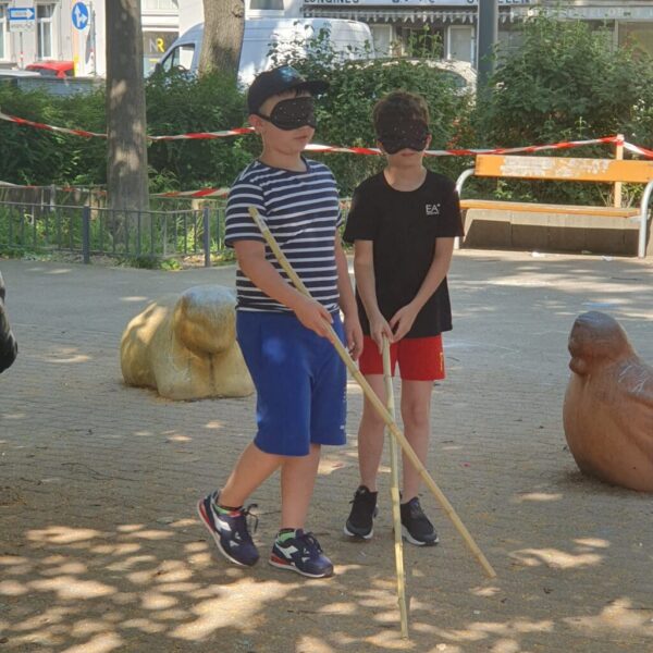 Es sind zwei Kinder im Park zu sehen, die dunkle Augenbinden am Kopf tragen und einen Holzstock in der Hand halten.