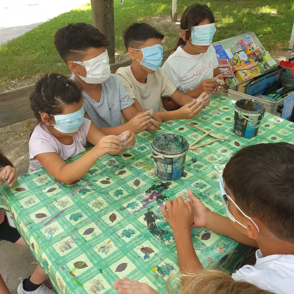 Fünf Kinder sitzen im Park um einen Tisch. Jedes trägt eine Augenbinde und hält einen Klumpen Ton in der Hand.