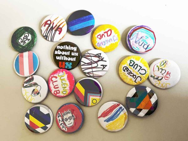 Fünfzehn Buttons, die verschieden gestaltet sind. Auf manche sind Namen wie zum Beispiel Aya, Andi und Jose zu lesen, manche sind mit der LGBTIQ+-Fahne gestaltet.