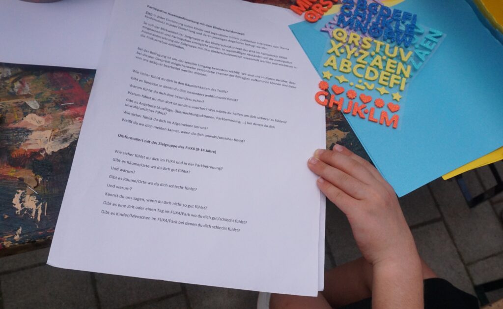 Ein Kind sitzt an einem Tisch und hält ein weißes Blatt Papier mit gedrucktem Text in der Hand. Der Text enthält Fragen zur partizipativen Auseinandersetzung mit dem Kinderschutzkonzept, speziell für Kinder im Alter von 9 bis 14 Jahren.
