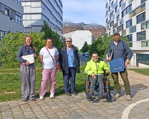 Vier Personen stehen auf einem Weg aus Kopfsteinpflaster, eine sitzt im Rollstuhl. Links und rechts sind Wohnhäuser zu sehen, im Hintergrund sieht man zwei Gasometer.