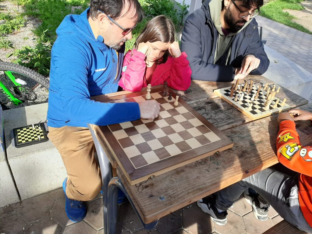 Vier Personen sitzen an einem Tisch, auf dem zwei  Schachbretter liegen.  Jeweils zwei Personen sind an einem Brett beschäftigt. Links sind nur zwei weiße und zwei schwarze Figuren am Brett, rechts ist Schachpartie noch nicht sehr weit fortgeschritten.