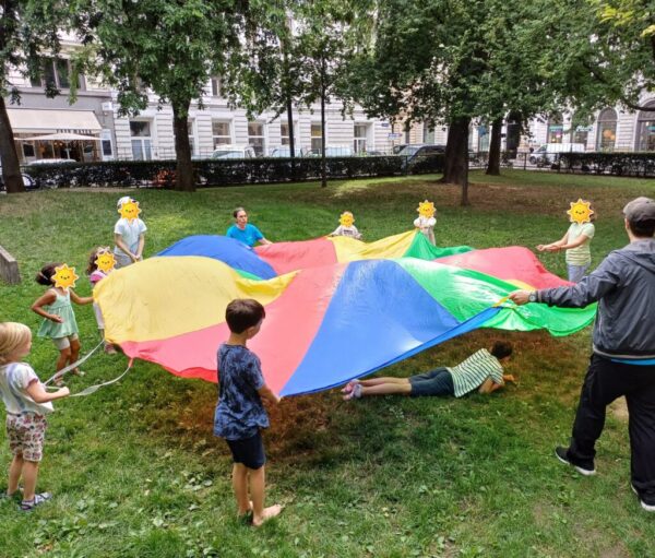Eine Gruppe von Menschen, vor allem Kinder, stehen auf einer Wiese unter Bäumen und halten ein Schwungtuch.