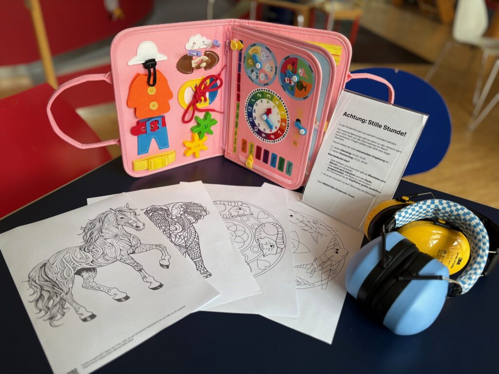 Ein Tisch, auf den ein blauer und ein gelber geräuschdämmender Kopfhörer und mehrere Ausmalbilder liegen, ein Fidget-Buch und ein Ständer mit einem Erklär-Text zur Stillen Stunde steht.