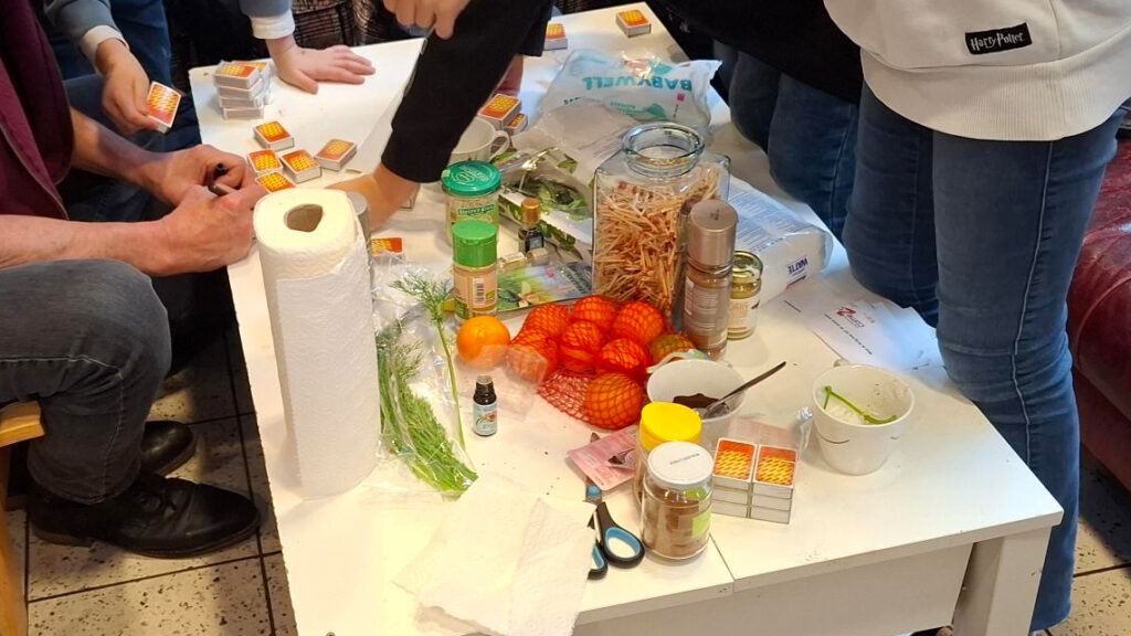 Auf einem Tisch liegen verschieden Gewürze, Duftöle, ein Sack Mandarinen, Streichhölzer und Streichholzschachteln. Im Vordergrund steht eine Küchenrolle. Es stehen einige Personen um den Tisch herum.