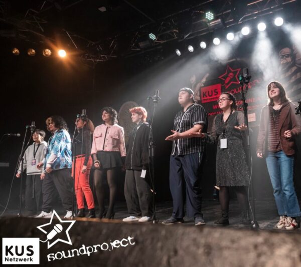 Gemini said Eine Gruppe von acht Jugendlichen steht nebeneinander auf einer beleuchteten Bühne und singt gemeinsam in Mikrofone. Im Hintergrund ist ein Banner mit der Schrift „KUS Soundproject“.