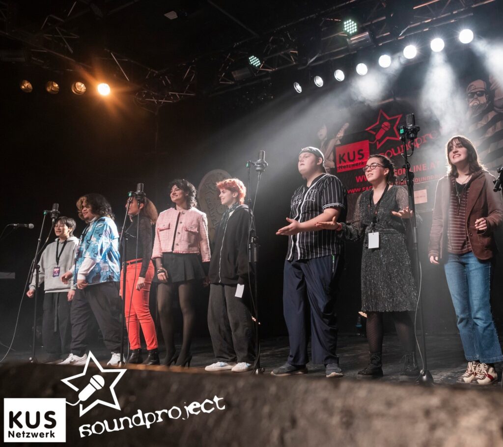 Gemini said Eine Gruppe von acht Jugendlichen steht nebeneinander auf einer beleuchteten Bühne und singt gemeinsam in Mikrofone. Im Hintergrund ist ein Banner mit der Schrift „KUS Soundproject“.