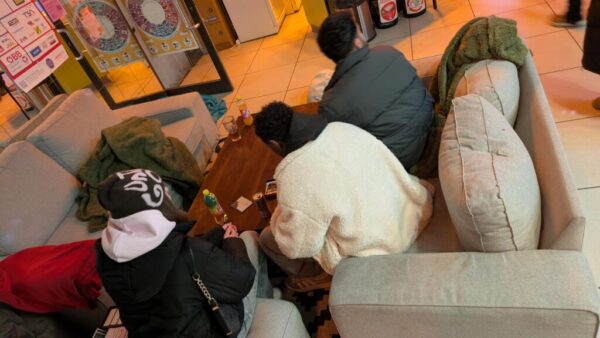 Drei Personen sitzen auf einer grauen Couchlandschaft rund um einen Couchtisch.