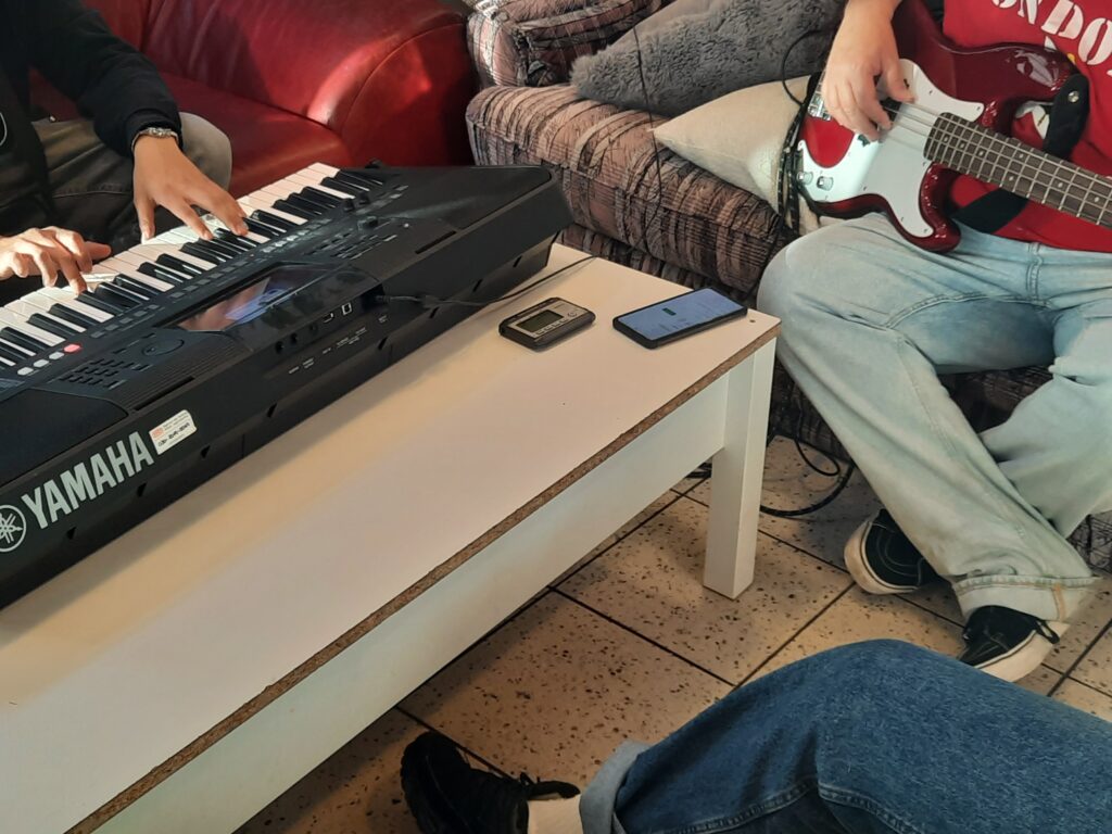 Zwei Personen machen gemeinsam Musik im Sitzen: Links spielt jemand auf einem Yamaha-Keyboard, rechts spielt eine Person auf einem roten E-Bass.