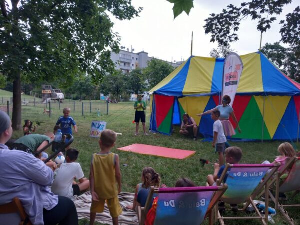 Kinder-Mitmachzirkus auf einer Wiese im Park. Im Hintergrund steht ein großes, buntes Zirkuszelt in Blau, Gelb und Rot. Im Vordergrund schauen Kinder und Erwachsene auf Stühlen und Picknickdecken zwei verkleideten Animateuren bei der Aufführung zu.