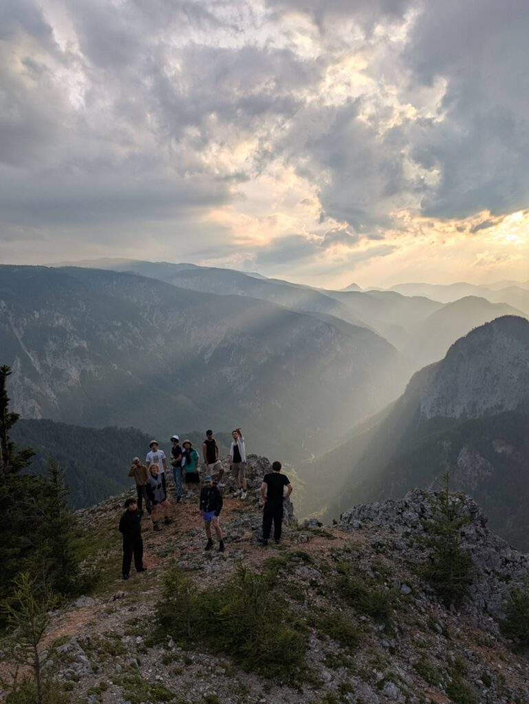 Mehrere Jugendliche stehen auf einem felsigen Berg. Hinter ihnen sind hohe Berge und ein tiefes Tal mit Wald. Durch die Wolken scheint die Sonne und schickt helle Strahlen ins Tal.
